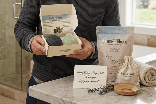Father holding an Onsen & Bloom Konjac Sponge Duo gift box with a Serenity shower steamer pouch nearby, in a calm modern bathroom