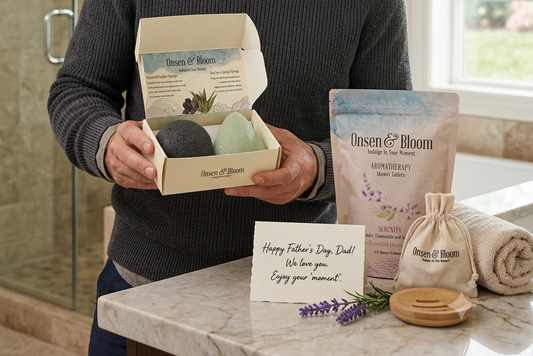 Father holding an Onsen & Bloom Konjac Sponge Duo gift box with a Serenity shower steamer pouch nearby, in a calm modern bathroom